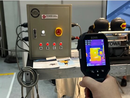 A thermal camera held up in front of an electrical control box, its screen showing the same box as a thermal image